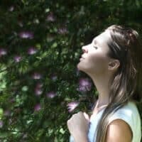 O femeie într-o cămașă albă stă cu ochii închiși, cu fața la soare, înconjurată de frunziș verde și flori violet, îmbrățișând energia de întinerire a suplimente naturale a naturii.